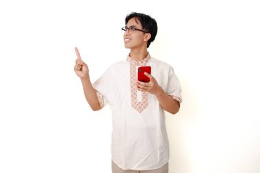 Geleneksel Müslüman gömlekli, kendine güvenen Endonezyalı adam elinde akıllı telefon tutarken beyaz arka planı işaret ediyor.