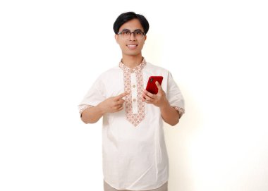 Geleneksel Müslüman gömlekli, kendine güvenen Endonezyalı adam elinde akıllı telefon tutarken beyaz arka planı işaret ediyor.