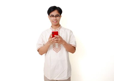 Geleneksel Müslüman gömlekli gülümseyen Endonezyalı adam, beyaz arka planda izole edilmiş ve cep telefonu kullanıyor.