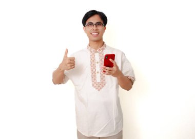 Geleneksel Müslüman gömlekli, kendine güveni tam bir Endonezyalı. Elinde akıllı telefon, beyaz arka planda izole bir şekilde baş parmak hareketi yapıyor.