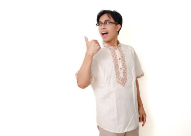 Geleneksel Müslüman gömlekli mutlu Endonezyalı adam temiz beyaz arka planda izole edilmiş, baş parmağıyla ayakta duruyor ve kenarı işaret ediyor.
