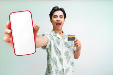 Yazlık gömlekli mutlu Asyalı adam elinde kredi kartı, online ödeme konsepti, rezervasyon ve mobil uygulama reklamı tutarken akıllı telefon modeli ekranı gösteriyor..