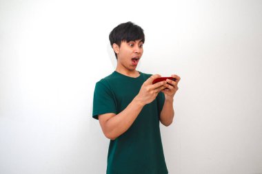 Beyaz arka planda izole edilmiş zafer jesti gösterirken akıllı telefondan mobil oyun oynayan mutlu Asyalı genç adam. Reklam ve tanıtım için mükemmel.