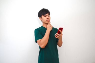 Genç Asyalı bir adam akıllı telefonuna bakarken düşünceli görünür, bir çözüm düşünürken ya da yeni bir fikir edinirken. Temiz, beyaz bir arkaplanı olan yeşil bir tişört giyiyor.