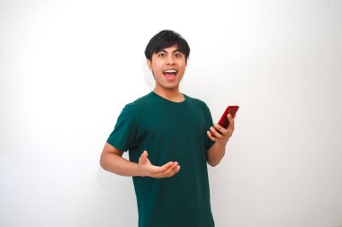 Genç Asyalı bir adam akıllı telefonundan bir şeye bakarken şaşırmış ve mutlu görünüyor. Yeşil tişörtlü adam düz beyaz bir arka planın önünde duruyor.