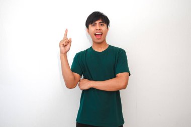 Genç Asyalı bir adam ağzı açık şaşırmış görünüyor ve parmağını havaya kaldırıyor, görünüşe göre harika bir fikri var. Beyaz arka planda koyu yeşil bir tişört giyiyor.