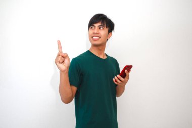 Mutlu bir Asyalı genç adam akıllı telefonu tutar ve gülümseyen bir ifadeyle yukarı doğru işaret eder, bir fikir veya çözüm önerir. Temiz, beyaz bir arkaplanı olan yeşil bir tişört giyiyor.