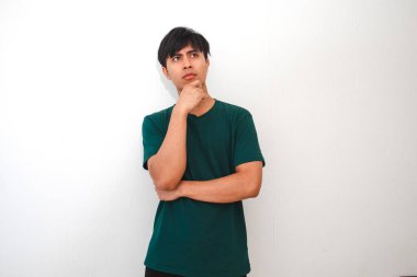 Genç bir Asyalı adam düşünceli bir ifadeyle yukarı ve yana bakıyor, eli çenesinde, sanki bir şey düşünüyormuş gibi. Düz beyaz bir arka planda koyu yeşil bir tişört giyiyor.