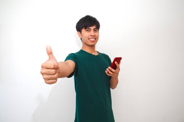 Akıllı telefon tutarken gülümseyen mutlu Asyalı bir genç kameraya olumlu bir jest yapıyor, beyaz arka planda izole edilmiş, terfi ve reklam için mükemmel.