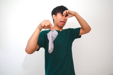 İzole edilmiş beyaz arka planda pis kokulu çoraplar tutan Asyalı genç adam, reklam için stüdyo yaşam tarzı konseptinde komik bir tepki gösteriyor.