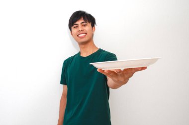 Kameraya boş bir yemek tabağı gösterirken gülümseyen genç Asyalı adam, beyaz arka planda izole edilmiş, yemek reklamları, promosyonlar veya restoran kampanyaları için mükemmel.