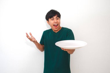 Asyalı genç bir adam, izole edilmiş beyaz arka planda şok olmuş bir yüzle boş bir yemek tabağına bakıyor. Yaşam tarzı veya reklam konsepti için bir stüdyo ortamında şaşkınlığını dile getiriyor.