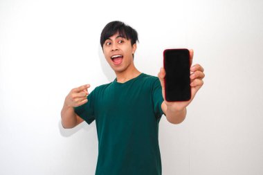 Akıllı telefonu boş ekranla gösteren mutlu Asyalı genç adam izole edilmiş beyaz arka planda heyecanla gülümsüyor. Teknoloji, promosyon ve reklam temaları için mükemmel.