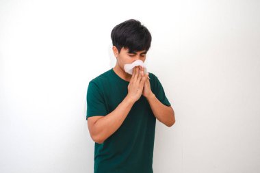 Genç Asyalı bir adam burnunu ve ağzını kapatırken mendile hapşırıyor, temiz beyaz bir arka planda izole edilmiş. Sağlık, hijyen ya da reklam kavramları için mükemmel.