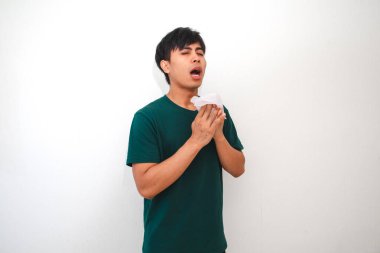 Genç Asyalı bir adam burnunu ve ağzını kapatırken mendile hapşırıyor, temiz beyaz bir arka planda izole edilmiş. Sağlık, hijyen ya da reklam kavramları için mükemmel.