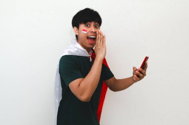 Elinde bayrak ve akıllı telefon olan heyecanlı Endonezyalı bir adam, beyaz arka planda izole edilmiş, ulusal kutlama ve reklam kavramları için mükemmel bir jest yapıyor..