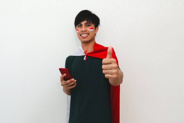 Endonezyalı adam elinde ulusal bayrak ve cep telefonu tutarken gülümsüyor, kutlama veya reklam için izole edilmiş beyaz arka planda mutlu yüzüyle kameraya başparmaklarını gösteriyor..