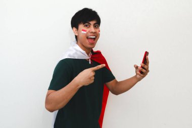 Heyecanlı Endonezyalı adam elinde bir bayrak tutuyor ve beyaz izole edilmiş arka planda mutlu bir ifadeyle akıllı telefonu işaret ediyor, bağımsızlık kutlaması ve reklam için mükemmel..