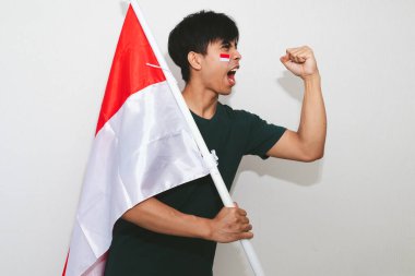 Heyecanlı genç Asyalı Endonezyalı bir destekçi olarak Endonezya bayrağını tutuyor ve beyaz arka planda izole edilmiş yumruklarıyla bağırıyor.
