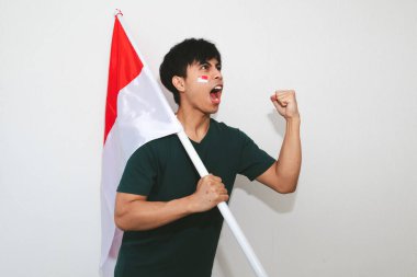 Heyecanlı genç Asyalı Endonezyalı bir destekçi olarak Endonezya bayrağını tutuyor ve beyaz arka planda izole edilmiş yumruklarıyla bağırıyor.