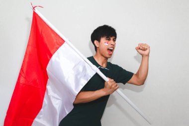 Heyecanlı genç Asyalı Endonezyalı bir destekçi olarak Endonezya bayrağını tutuyor ve beyaz arka planda izole edilmiş yumruklarıyla bağırıyor.