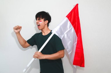 Heyecanlı genç Asyalı Endonezyalı bir destekçi olarak Endonezya bayrağını tutuyor ve beyaz arka planda izole edilmiş yumruklarıyla bağırıyor.