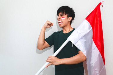 Heyecanlı genç Asyalı Endonezyalı bir destekçi olarak Endonezya bayrağını tutuyor ve beyaz arka planda izole edilmiş yumruklarıyla bağırıyor.