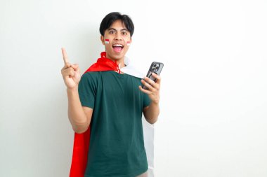 Heyecanlı genç Endonezyalı adam ulusal bayrağı elinde tutarak Bağımsızlık Günü 'nü kutluyor ve beyaz bir arka planda izole edilmiş bir şekilde kenarı işaret ederken akıllı telefon kullanıyor.