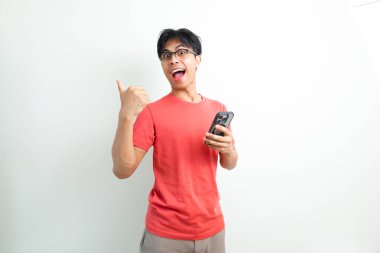 Turuncu tişört giyen neşeli genç Asyalı adam elinde akıllı telefon tutarken ve başparmağıyla işaret ederken beyaz izole edilmiş arka planda komik heyecanlı bir yüz ifadesi takınıyor.