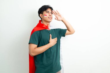 Endonezyalı gururlu genç bir adam Endonezya bayrağını tutuyor ve el ele selamlıyor, Bağımsızlık Günü 'nü parlak bir arka planda vatansever bir ifadeyle kutluyor.