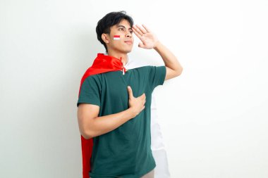 Endonezyalı gururlu genç bir adam Endonezya bayrağını tutuyor ve el ele selamlıyor, Bağımsızlık Günü 'nü parlak bir arka planda vatansever bir ifadeyle kutluyor.