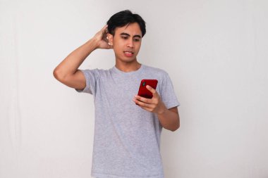 Beyaz arka planda izole edilmiş akıllı telefona bakarken kafası karışmış genç Asyalı bir adam kafasını kaşıyordu. Teknoloji sorunları, çevrimiçi sorunlar ya da başarısız bağlantı kavramları için mükemmel.