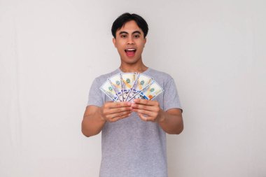 Asyalı mutlu genç adam ayakta duruyor ve elinde heyecanlı bir ifadeyle bir sürü para tutuyor, beyaz arka planda soyutlanmış.