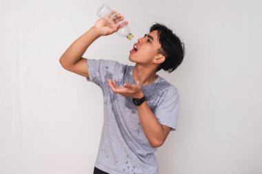 Asyalı yorgun genç bir adam beyaz arka planda izole edilmiş hayal kırıklığına uğramış bir ifadeyle terliyor ve elinde boş bir su şişesi tutuyordu. Susuzluk, sıcaklık ya da susuzluk kavramı