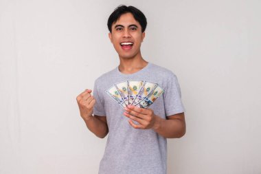 Kutlamada elinde para tutan ve yumruğunu sıkan mutlu Asyalı genç bir adamın portresi, izole edilmiş beyaz bir arka plana karşı neşeyle gülümsüyor, reklam için mükemmel.
