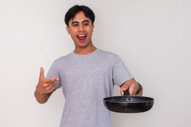 Heyecanlı genç Asyalı adam elindeki boş bir kızartma tavasını işaret ederek izole edilmiş beyaz arka planda mutlu bir yüz ifadesi gösteriyor.