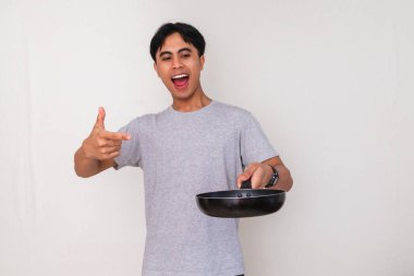 Heyecanlı genç Asyalı adam elindeki boş bir kızartma tavasını işaret ederek izole edilmiş beyaz arka planda mutlu bir yüz ifadesi gösteriyor.