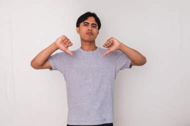 Kızgın genç Asyalı adam, beyaz arka planda duran, çılgın bir ifadeyle baş parmak hareketi yapıyor. Reddetme, protesto ya da olumsuz geri bildirim konsepti için mükemmel