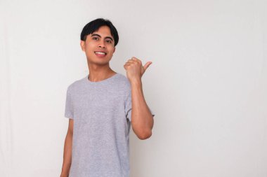 Gülümseyen genç bir Asyalı 'nın portresi beyaz izole bir arka planda başparmağıyla yan tarafı işaret ediyor. Reklam, promosyon ve sunum modeli için mükemmel.