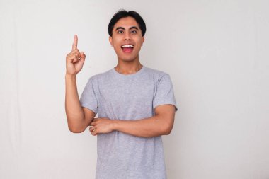Heyecanlı genç Asyalı adam, sanki beyaz arka planda izole edilmiş yeni bir fikir edinmiş gibi ilham verici bir ifadeyle işaret ediyor. Yaratıcı, yenilikçi veya promosyon kavramları için mükemmel.