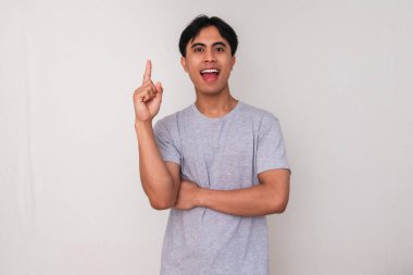 Heyecanlı genç Asyalı adam, sanki beyaz arka planda izole edilmiş yeni bir fikir edinmiş gibi ilham verici bir ifadeyle işaret ediyor. Yaratıcı, yenilikçi veya promosyon kavramları için mükemmel.