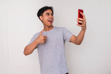 Beyaz arka planda izole edilmiş akıllı bir telefonla selfie ya da video çekerken gülümseyen Asyalı genç bir adamın portresi