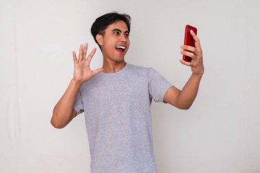 Beyaz arka planda izole edilmiş akıllı bir telefonla selfie ya da video çekerken gülümseyen Asyalı genç bir adamın portresi