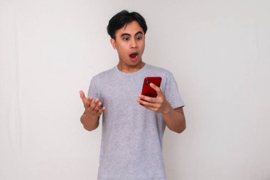 Beyaz arka planda izole edilmiş, şok olmuş ve ciddi bir ifadeyle akıllı telefonuna bakan Asyalı genç bir adamın portresi.