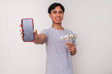 Elinde para tutan gülümseyen Güneydoğu Asyalı genç bir adamın portresi ve izole edilmiş beyaz bir arkaplanda kameraya boş bir akıllı telefon ekranı gösteriyor.