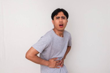 Komik genç Asyalı adam, sanki kabızlık çekiyormuş gibi acı dolu bir yüzle karnını tutuyor. Beyaz arkaplanda izole