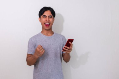 Heyecanlı Asyalı genç adam, beyaz arka planda izole bir şekilde bağırıp cep telefonu kullanarak başarıyı kutluyor.