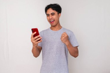 Heyecanlı Asyalı genç adam, beyaz arka planda izole bir şekilde bağırıp cep telefonu kullanarak başarıyı kutluyor.