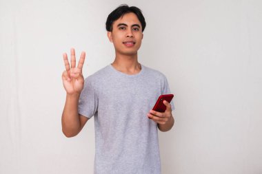 Akıllı telefon tutan gülümseyen Asyalı genç bir adamın portresi ve beyaz izole edilmiş arka planda üç parmak hareketi gösteriyor. Teknoloji, iletişim ve kampanya reklamları için mükemmel.