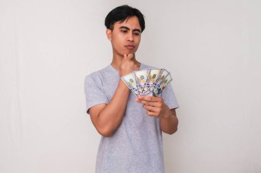Genç Asyalı bir adamın portresi, beyaz arka planda izole edilmiş, elindeki paraya bakarken düşünüyor. Finansal, ticari ve reklam konuları için mükemmel